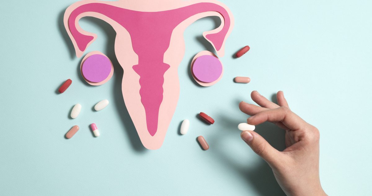 Paper cutout of uterus with surrounding pills and hand holding white pill